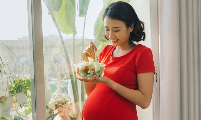 Young pregnant woman eating vegetable salad near window at home